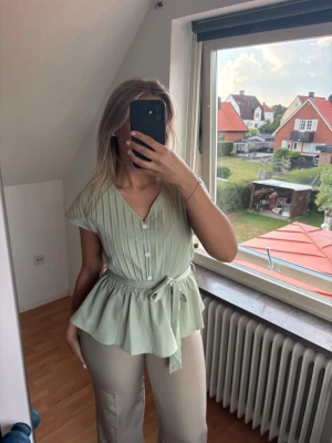 Ljusgrön blus med knyt och knappar - Säljer en ljusgrön blus med v-ringning, knappar framtill och ett snyggt knyt i midjan. Blusen har korta ärmar och ett plisserat tyg som ger en extra touch. Perfekt för dig som gillar en feminin och trendig stil.