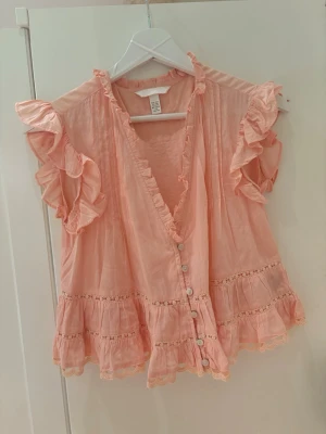 Persikofärgad blus med volanger H&M - Supersöt persikofärgad blus från H&M med knappar framtill, volangdetaljer längs ärmar och nederkant samt spetsinslag. Blusen har v-ringning och korta, ryschiga ärmar. Perfekt för en romantisk och trendig look.
