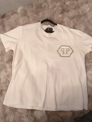 Vit t-shirt med strassdetaljer qp - Säljer en vit t-shirt med korta ärmar och ett stort PP-logotypmotiv i strass på bröstet. T-shirten har en klassisk passform och är gjord i mjuk bomull, perfekt för en clean och stilren look.