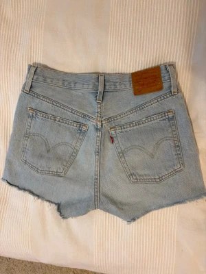 Ljusblå jeansshorts 501’ från Levi's - Säljer ett par klassiska ljusblå jeansshorts från Levi's med rå kant nertill och femficksmodell. Shortsen har misrise och är tillverkade i bomull. Perfekta för varma dagar och har den ikoniska Levi's-lappen bak. Brukar i vanliga fall ha stl. W27.