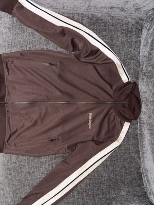 Brun hoodie från Palm Angels - Säljer en brun hoodie från Palm Angels med dragkedja framtill och vita samt svarta ränder längs ärmarna. Hoodien har två fickor med dragkedja och Palm Angels-logga broderad på bröstet. Tillverkad i ett mjukt och stretchigt material som känns sportigt.