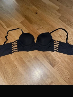 Svart push-up bh med banddetaljer - Snygg svart push-up bh med vadderade kupor och flera tunna band på sidorna som ger en trendig look. Axelbanden är justerbara och bh:n har en slät finish i ett mjukt material. Perfekt för dig som vill ha något extra under kläderna.