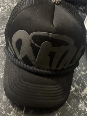 Svart truckerkeps med tryck - Snygg svart truckerkeps med stort matt svart tryck framtill och dekorativt snöre ovanför skärmen. Baksidan är i mesh för extra ventilation och kepsen har en liten metallogo på toppen. Perfekt för en streetwear-look.