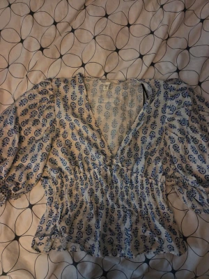 Blommig blåvit blus med puffärm - Söt blus i vitt med blått blommönster och puffiga långärmar. Djup v-ringning och smockad midja som ger en snygg siluett. Tillverkad i ett lätt och luftigt tyg, perfekt för en trendig och avslappnad look.