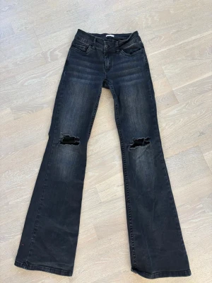 Svarta bootcut jeans från Nelly - Snygga svarta bootcut jeans från Nelly med slitningar över knäna. Jeansen har klassisk femficksdesign, hög midja och utsvängda ben. Perfekta för dig som gillar en trendig och avslappnad stil.Lite slitna nertill på höger sida se bild.