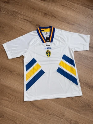 Sverige Adidas Vm94 Matchtröja S - Vit svensk landslagströja från Adidas med blå och gula detaljer på framsidan, broderad svensk sköld och Adidas-logga. Kragen är blå, gul och röd med V-ringning. Tillverkad i lätt och ventilerande material, perfekt för sport och träning.