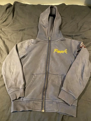 Grå zip-hoodie från Peak Performance - Säljer en grå hoodie med dragkedja från Peak Performance. Hoodien har huva, fickor framtill och gul brodyr med texten 'Peak' på bröstet. På ena ärmen finns ett snyggt broderat märke med logga. Perfekt för chill dagar eller till skolan.