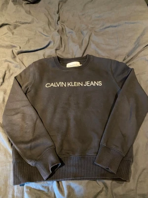 Svart sweatshirt från Calvin Klein Jeans - Svart sweatshirt från Calvin Klein Jeans med vit logga tryckt på bröstet. Klassisk rund halsringning och ribbade muddar vid ärmslut och nederkant. Perfekt för en stilren och avslappnad look.