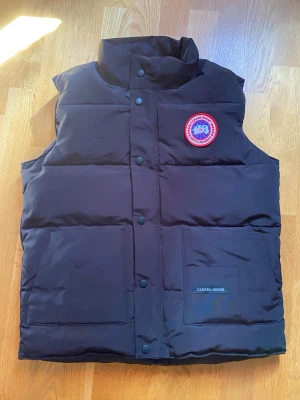 Svart dunväst från Canada Goose - Snygg svart dunväst från Canada Goose med klassisk logga på bröstet och hög krage. Västen har tryckknappar framtill och två stora fickor. Perfekt för lager-på-lager och håller dig varm under kyliga dagar.
