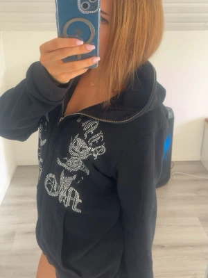 Svart hoodie med strassmotiv från ”Aim for the moon” - Säljer en svart hoodie med dragkedja och huva. Framsidan är dekorerad med stora strassmotiv och text i glittrande stenar. Hoodien har långa ärmar och en avslappnad passform, perfekt för en trendig och bekväm look.
