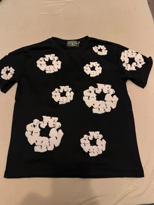 Svart t-shirt med vita grafiska tryck - Snygg svart t-shirt med unika vita grafiska tryck i cirkelformade mönster över hela tröjan, både fram och bak. T-shirten har rund hals och korta ärmar. Perfekt för dig som vill sticka ut med en cool och modern stil.