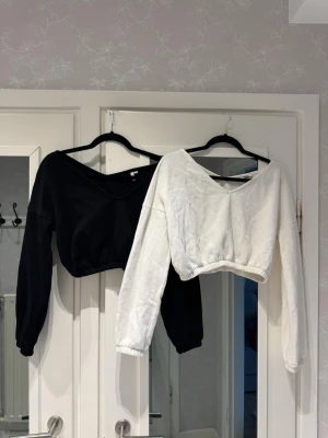 2 croppade tröjor från NLY Trend XS - Två croppade långärmade tröjor från NLY Trend i storlek XS. En är svart och en är vit, båda med bred v-ringning och resår i midjan. Säljs i duopaket