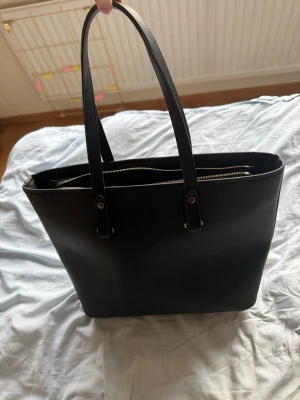 Svart tote handväska i läderimitation - Stilren svart tote handväska med långa handtag och dragkedja upptill. Väskan har en rymlig insida och är tillverkad i slät läderimitation. Perfekt för dig som vill ha plats för både laptop och vardagsprylar. Enkel och klassisk design som passar till det mesta.