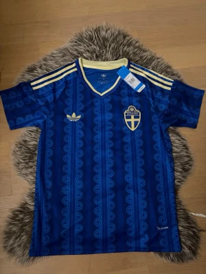 Blå Sverige fotbollströja Adidas - Säljer en blå Sverige fotbollströja från Adidas med gulddetaljer och retroinspirerat mönster. Tröjan har broderat landslagsemblem och Adidas-logga, samt gula ränder på axlarna och v-ringad krage. Perfekt för dig som älskar fotboll och vill sticka ut på läktaren. Helt ny,  sveriges 2026 borta tröja.