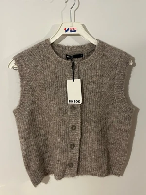 Beige stickad väst från Bik Bok - Mysig beige stickad väst från Bik Bok med ribbad struktur och knappar framtill. Perfekt att slänga över en skjorta eller t-shirt för en trendig lager-på-lager-look. Västen har en rund halsringning och är något croppad i modellen. Storlek S men passar även M. helt ny med prislapp, nypris 399:- pris kan diskuteras vid snabb affär