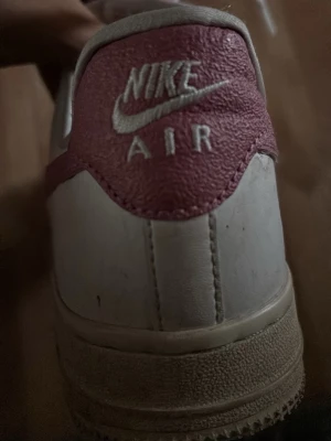 Nike Air Force 1 vita och rosa sneakers - Nike Air Force 1 sneakers i vitt läder med rosa detaljer och klassisk swoosh på sidan. Skon har perforerad tå för ventilation, platt sula och snörning framtill. Broderad Nike Air-logga bak i vitt på rosa bakparti. Perfekt för dig som gillar streetstyle.(Pris kan disskuteras)