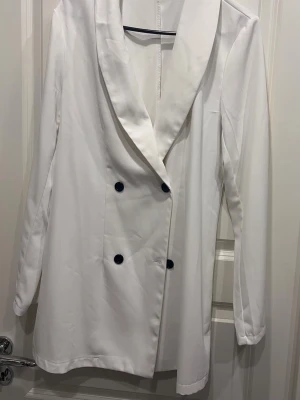 Vit dubbelknäppt kavaj - Stilren vit kavaj med dubbelknäppning och svarta knappar. Kavajen har lång ärm, bred krage och rak passform. Perfekt för dig som vill ha en klassisk och trendig look. Materialet ser ut att vara slätt, tunnt och något glansigt.