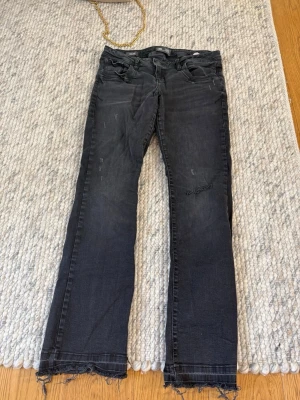Svarta bootcut jeans från LTB - Säljer ett par svarta bootcut jeans från LTB med slitna detaljer och råa kanter vid bensluten. Även gjort ett hål på ena knät. Jeansen har fem fickor och en klassisk knappgylf. Perfekta för dig som gillar en avslappnad och trendig stil. 