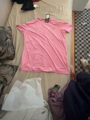 Rosa t-shirt från Under Armour - Säljer en rosa t-shirt från Under Armour med klassisk passform och korta ärmar. T-shirten har en diskret logga på bröstet och är tillverkad i ett mjukt, lätt material som passar perfekt till vardags eller träning.