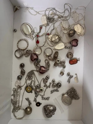 Blandat smyckespaket i silverton - Säljer ett mixat paket med smycken i silverfärg, inklusive halsband, ringar, örhängen och berlocker. Det finns detaljer som hjärtan, löv, blommor och stenar i lila och rött. Materialet är mestadels metall med inslag av glas och plast. Vissa smycken kan ha silver men ej 100% då de ej är noggrant sökta.