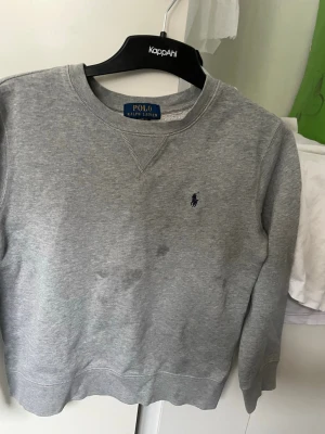 Grå sweatshirt från Polo Ralph Lauren - Klassisk grå sweatshirt från Polo Ralph Lauren med rund halsringning och broderad marinblå logga på bröstet. Tillverkad i mjuk bomullsblandning och har ribbade muddar vid ärmslut och nederkant. Perfekt för en avslappnad och stilren look. Fläckarna kan gå bort men eftersom den är för liten för mig pallar jag inte att tvätta den. Ingen slitage 