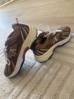 Bruna chunky sneakers med vita detaljer - Säljer ett par bruna chunky sneakers med vita och vinröda detaljer. Skorna har meshpaneler för extra ventilation, vita snören och en tjock vit sula med svarta och vinröda inslag. Perfekta för en sportig och trendig look.