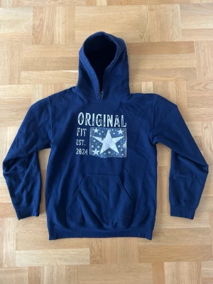 Original hoodie - Mörkblå hoodie med stor huva och magficka. Tryck på bröstet med texten 'ORIGINAL FIT EST. 2024' och en stor vit stjärna omgiven av mindre stjärnor. Perfekt för en avslappnad och cool stil. Mjuk bomullskänsla. Limiterad och går ej att köpa längre!