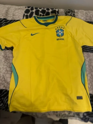 Gul Brasilien fotbollströja Nike - Säljer en klassisk gul Brasilien fotbollströja från Nike med gröna och blå detaljer. Tröjan har CBF-emblemet och fem stjärnor broderat på bröstet samt Nike-logga. Perfekt för fotbollsfans som vill visa sitt stöd för landslaget.