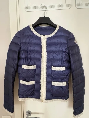 Mörkblå moncler - Snygg mörkblå dunjacka med fyra framfickor och vita volangdetaljer längs kanter, halsringning och fickor. Västen har quiltad design och rund hals. Perfekt för dig som vill ha en unik och trendig look.