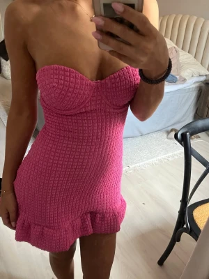 Rosa axelbandslös kort klänning - Säljer en superfin rosa kort klänning med axelbandslös design och markerad byst. Klänningen har en ribbad struktur och volangkant nertill som ger en söt touch. Perfekt för sommarens alla tillfällen!