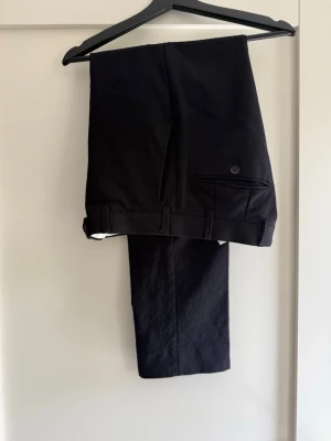 Svarta kostymbyxor med fickor - Snygga svarta kostymbyxor med klassisk rak passform och bakficka med knapp. Byxorna har hällor för bälte och är tillverkade i ett slätt tyg som ger en stilren look. Perfekta för dig som vill ha en clean och dressad stil.