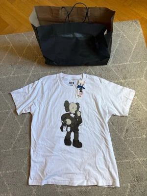 Vit KAWS t-shirt  - Säljer en vit t-shirt från  KAWS med en stor grafisk print av KAWS-figuren på framsidan. T-shirten är kortärmad och gjord i mjuk bomull, perfekt för en streetwear-inspirerad look. Köpte tröjan i USA för 100 dollar typ 1000kr, köpt lite utanför New Jersey i USA. Aldrig använd och är helt ny. Påse ingår såklart. 