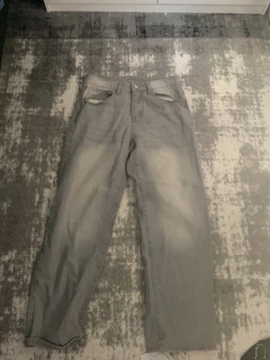 Grå raka jeans med slitningar - Säljer ett par grå jeans med raka ben och diskreta slitningar vid fickorna. Jeansen har en klassisk femficksmodell och ljus tvätt med viss blekning på framsidan och baksidan. Perfekta för en avslappnad och trendig look.