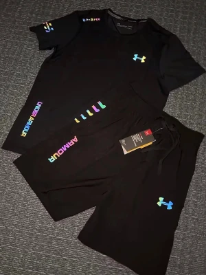 Svart träningsset från Under Armour - Säljer ett par svart träningsset från Under Armour med färgglada loggor och detaljer. Shortsen har snörning i midjan och är tillverkade i ett mjukt och lätt material som passar perfekt till träning eller avslappnade dagar. Storlek S, helt ny 