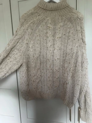 Beige kabelstickad polotröja - Säljer en chunky beige kabelstickad polotröja med breda ärmar och ribbade muddar. Tröjan har ett klassiskt flätmönster och hög krage som ger en mysig och trendig look. Perfekt för kyliga dagar och enkel att styla till olika outfits.