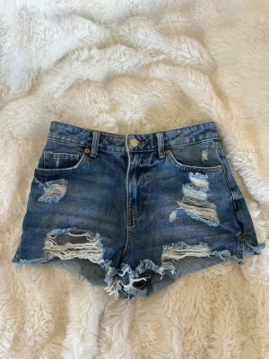 Blå slitna jeansshorts med fransar - Säljer ett par blå denimshorts med slitna detaljer och fransig kant. Shortsen har fem fickor, knappgylf och en klassisk midja. Perfekta för sommaren och ger en avslappnad vibe.