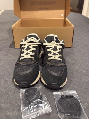 Svarta Polo Ralph Lauren sneakers - Snygga svarta sneakers från Polo Ralph Lauren med vita detaljer och vita skosnören. Skorna har en klassisk låg siluett, mesh och mocka-paneler samt Polo-logga på plösen. Två extra par svarta skosnören medföljer.