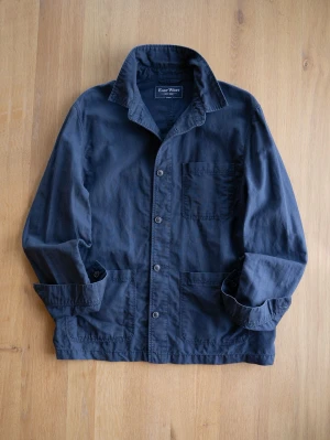Navy Chore Jacket, size M - Stilren mörkblå overshirt från East West med tre utanpåliggande fickor och knäppning framtill. Skjortkrage och raka ärmar ger en avslappnad look. Tillverkad i ett mjukt bomullstyg som passar perfekt till lager på lager.