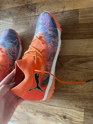 Orangea Puma fotbollsskor - Säljer ett par grymma orangea fotbollsskor från Puma med svart logga på sidan. Skorna har snörning, vit sula och ett mönstrat parti i blått och orange framtill. Ovandelen är i textilmaterial som ger bra andningsförmåga och passform.