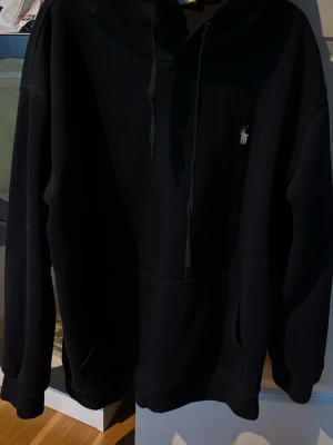 Svart hoodie från Polo Ralph Lauren - Klassisk svart hoodie från Polo Ralph Lauren med huva och dragsnören. Har en liten broderad logga på bröstet och en stor magficka framtill. Tillverkad i mjukt material som känns skönt mot huden. Perfekt för en avslappnad och stilren look. Obs - det är en L men jag skulle nog säga att den passar en M. 
