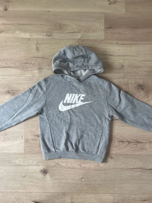 Grå hoodie från Nike med logga - Snygg grå hoodie från Nike med stor vit logga på bröstet. Hoodien har huva, ribbade muddar och magficka. Tillverkad i mjukt och bekvämt material, perfekt för en avslappnad stil. Passar dig som gillar sportigt och klassiskt.