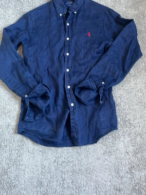 Marinblå linneskjorta Polo Ralph Lauren - Säljer en marinblå långärmad skjorta från Polo Ralph Lauren i slim fit. Skjortan är tillverkad i linne och har klassisk button-down krage, vita knappar och den ikoniska röda loggan broderad på bröstet.