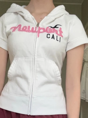 Vit hoodie med kort ärm Newport Cali - Vit hoodie med dragkedja och korta ärmar. Broderad text 'Newport' i rosa och 'CALI' i svart framtill samt en liten logga. Två fickor fram och huva. Säljer eftersom den var lite för kort( jag är 168). Tröjan har små fläckar som knappt syns på framsidan.
