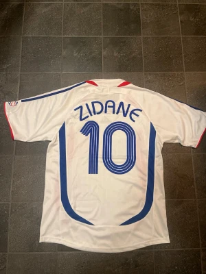 Frankrike 2006 - VM Zidane matchtröja - Säljer en vit matchtröja från Frankrikes landslag med Zidane och nummer 10 på ryggen. Tröjan har blå och röda detaljer, Adidas-logga och VM 2006-märke på bröstet. Korta ärmar och sportig passform, perfekt för fotbollsfans eller samlare. Är L men passar även M.