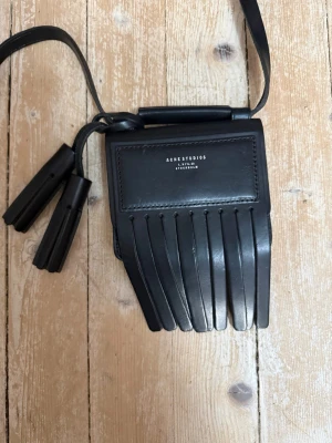 Svart fransad handväska Acne Studios -  Perfekt för dig som gillar en unik och trendig accessoar. Väska från Acne Studios i svart skinn med två svarta tofsar, i toppenskick!  Stängs enkelt med en magnet och rymmer det nödvändigaste. En liten innerficka finns för smått.  Mått: 15x12 cm (exklusive fransarna). OANVÄND. 
