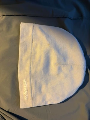Vit enkel mössa med broderad text - Säljer en stilren vit mössa i mjukt tyg med diskret broderad text på kanten. Mössan har en klassisk form och passar perfekt för dig som gillar minimalistisk stil. Materialet känns stretchigt och bekvämt.