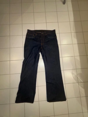 Mörkblå raka jeans med kontrastsömmar - Säljer ett par mörkblå jeans med bootcut ben och klassisk femficksdesign. Jeansen har kontrastsömmar i orange och detaljerade bakfickor. Tillverkade i kraftigt denimtyg som ger en snygg och tidlös look Nudiejeans.