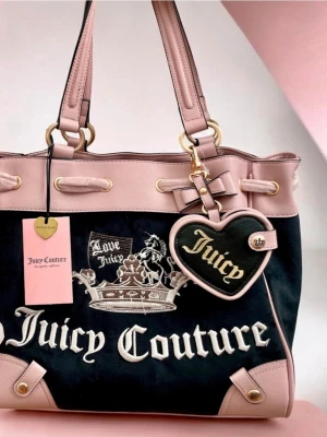 Juicy Couture svart och rosa handväska - Juicy Couture handväska i svart sammet med rosa detaljer i skinnimitation. Väskan har stora logotexter, guldfärgade metalldetaljer och ett hjärtformat nyckelband med texten 'Juicy'. Två axelband och dekorativ rosett på sidan.