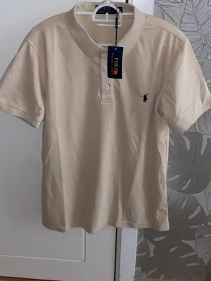 pikétröja från Polo Ralph Lauren - Klassisk beige pikétröja från Polo Ralph Lauren med korta ärmar och krage. Tröjan har knappar framtill och den ikoniska loggan broderad på bröstet. Perfekt till både jeans och shorts för en clean och stilren look.