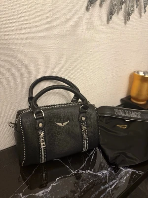 Svart handväska Zadig & Voltaire - Cool svart handväska i läder från Zadig & Voltaire med silverfärgade detaljer, dubbla handtag och dekorativa dragkedjor framtill. Väskan har en avtagbar axelrem med logga och kommer med tillhörande dustbag.